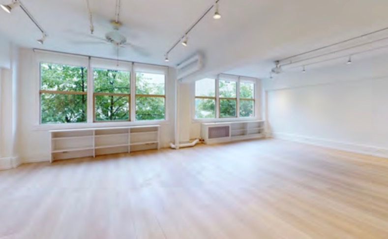 A corner of the studio with two sets of large wood-framed windows looking onto green trees, low white shelving beneath them, and a pale wood floor.