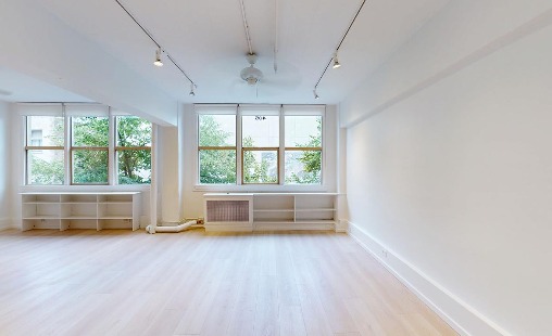 The main studio seen from the far wall: two banks of wood-framed windows with greenery, low white shelving, ceiling fan and track lights over a pale wood floor.